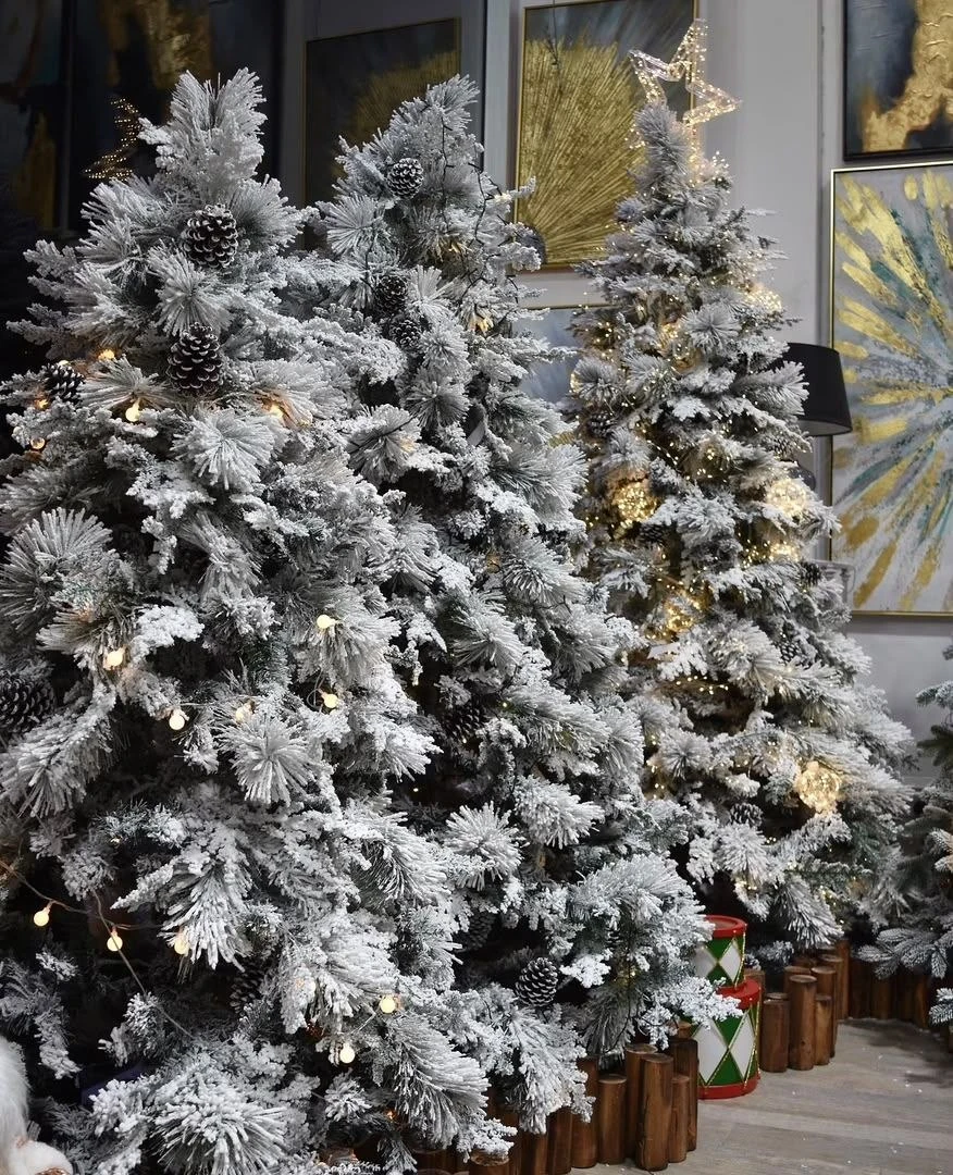 White flocking, pinecones and warm lights across artificial pine branches, with another decorated tree behind.